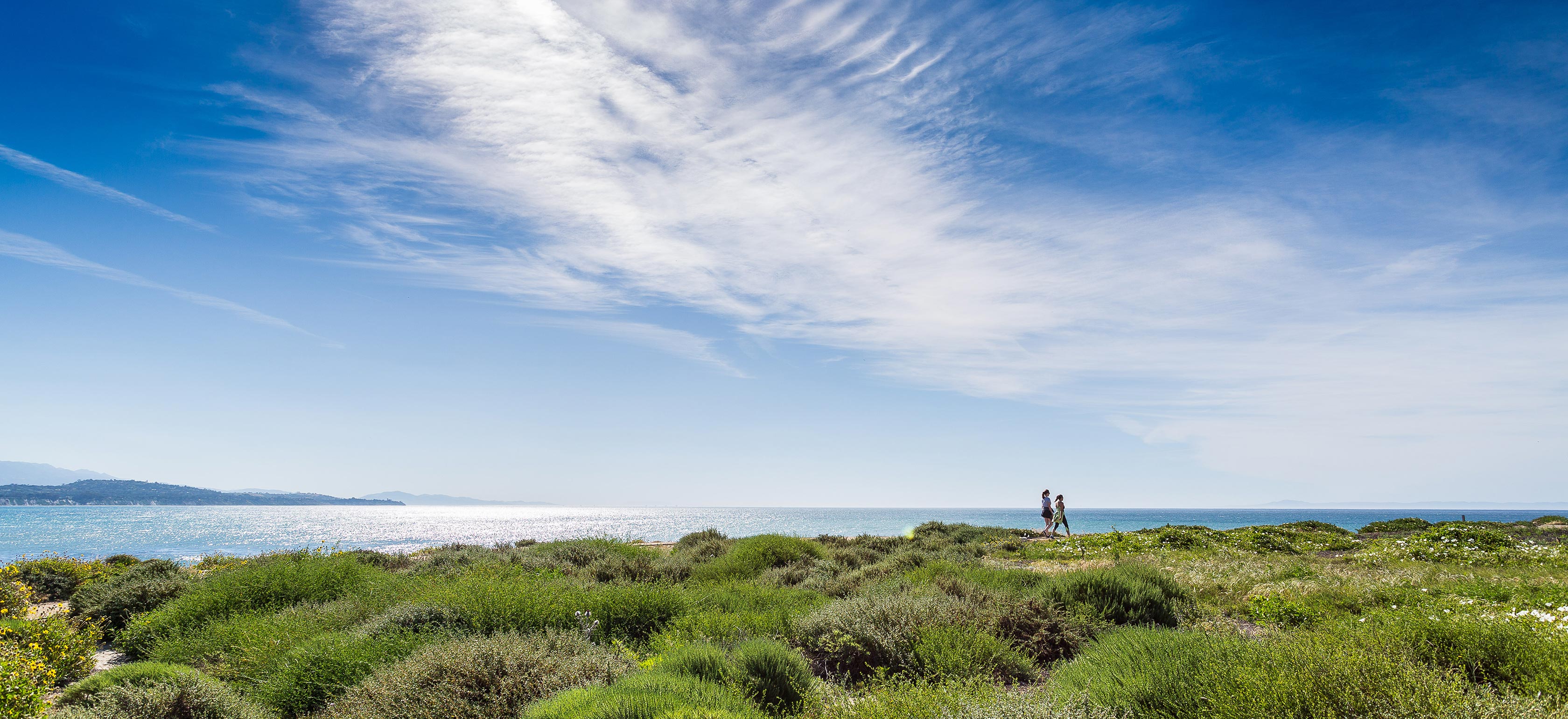 View of sky with streak of clouds with two people walking on cliff at the end of grassy area with ocean view 