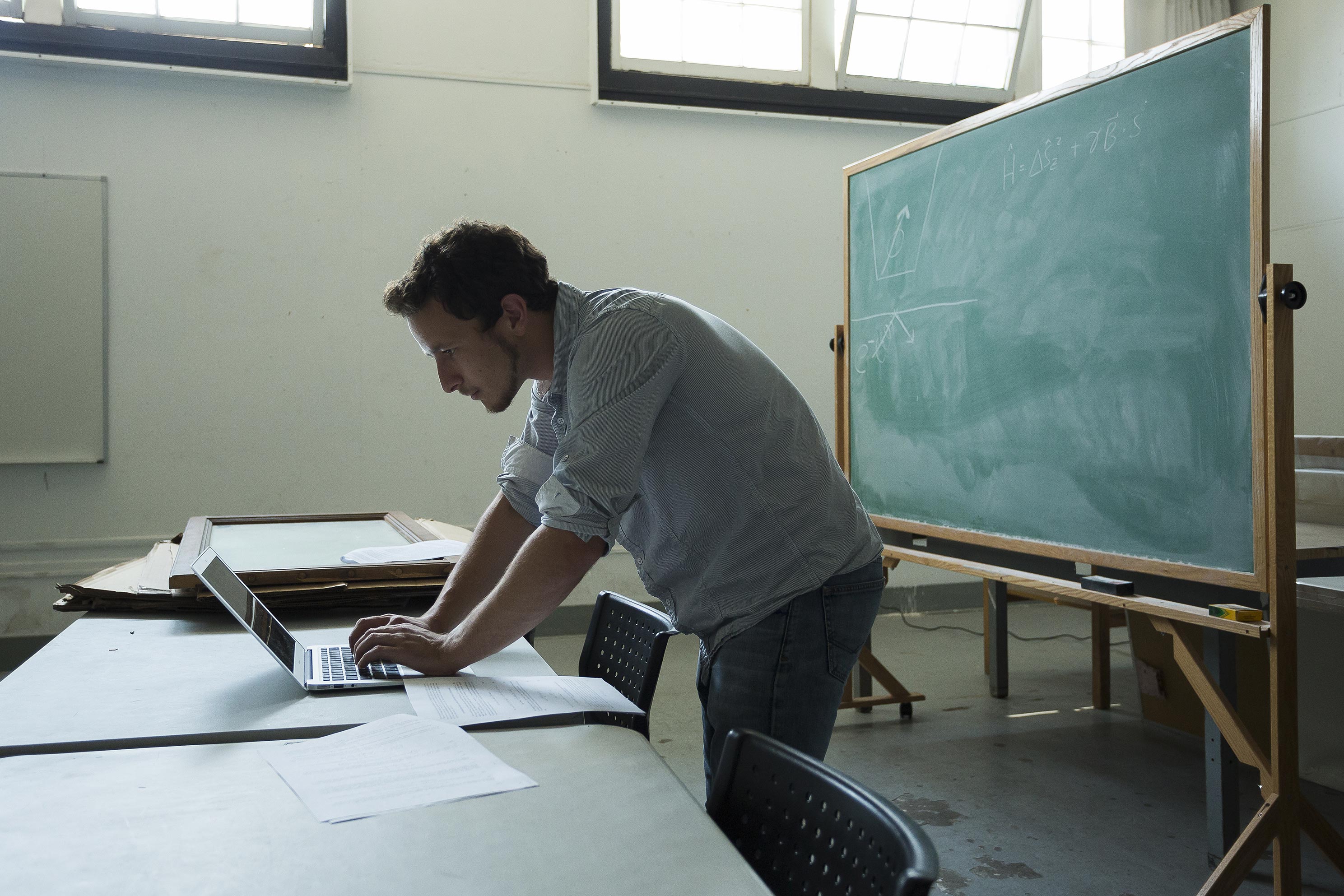 Student standing and typing on laptop with blackboard behind him