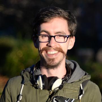 headshot of Jonathan Balkind from the shoulders up, wearing an olive green hooded jacket, black rimmed glasses, and smiling